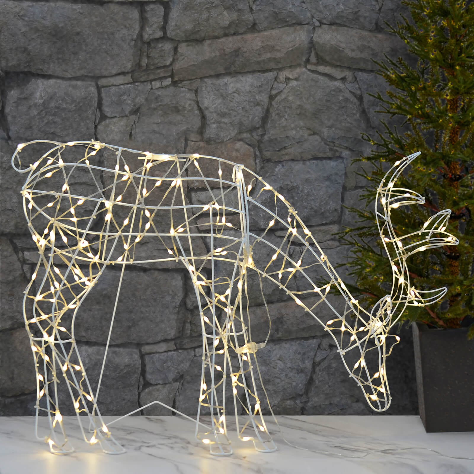Grazing reindeer outdoor Christmas decoration lit by warm white LED lights on a white wire frame, standing on a patio with stone wall and Christmas tree